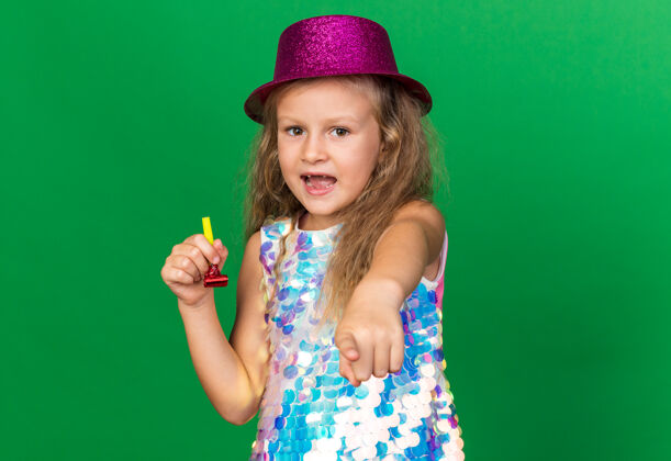 小一个惊讶的金发小女孩 戴着紫色的派对帽 手持派对哨子 孤立地指着绿色的墙壁 留着复制空间女孩指着金发