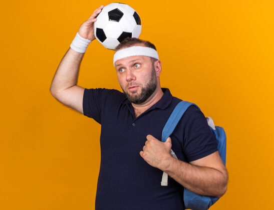 壁板惊讶的成年斯拉夫运动男子戴着背包头带和手环举行球在头上看一边孤立的橙色墙上复制空间头带穿成人