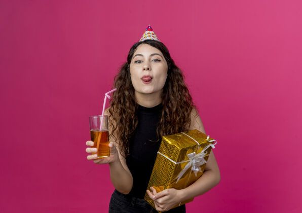 鸡尾酒年轻漂亮的女人 卷发 戴着节日帽 手里拿着生日礼盒和鸡尾酒 看着前面 快乐而积极地伸出舌头生日派对脸年轻盒子