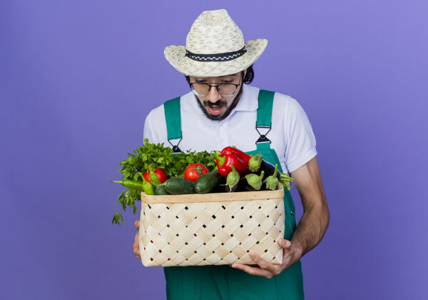 帽子年轻的留着胡须的园丁 穿着连体衣 戴着帽子 手里拿着装满蔬菜的板条箱 站在蓝色的墙上惊讶地看着它人姿势抱着