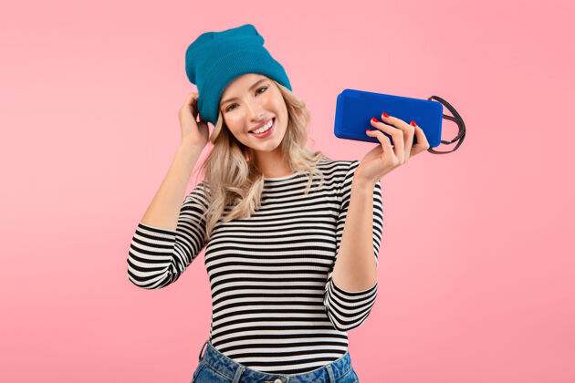 快乐年轻女子手持无线扬声器听音乐穿着条纹衬衫和蓝色帽子微笑着摆出粉红色的姿势情感女人服装