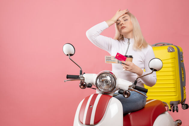 咖啡前视图疲惫的年轻女士坐在轻便摩托车上 手放在头上 手里拿着咖啡杯抱着轻便摩托车坐着
