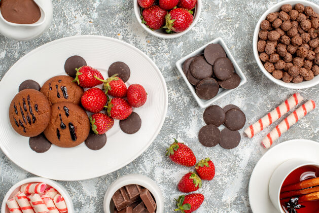 草莓顶近距离观察椭圆形盘子上的饼干草莓和圆形巧克力灰白色桌子上的糖果草莓巧克力麦片可可和肉桂茶碗食物肉桂谷类食品
