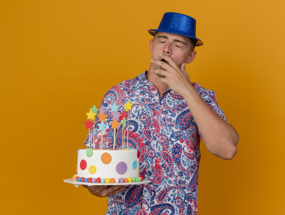 蛋糕打呵欠的年轻人戴着蓝色帽子拿着蛋糕孤立在橙色抱着帽子年轻人