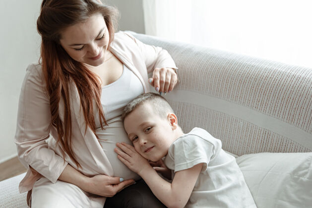 漂亮小男孩在家里的沙发上抱着怀孕母亲的肚子产妇期望父母