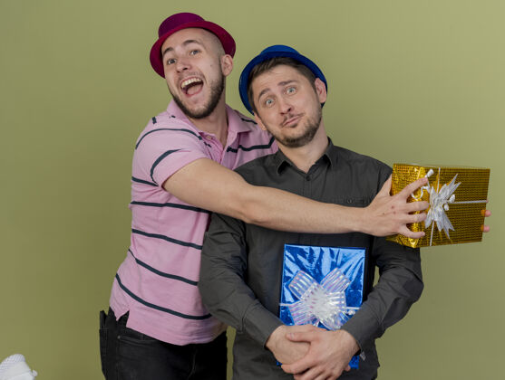 小伙子快乐的年轻人戴着红帽子拿着礼品盒 拥抱着令人印象深刻的家伙拿着礼品盒戴着蓝色帽子隔离在橄榄绿印象穿着年轻人