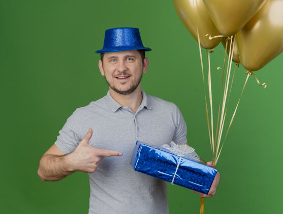 举行微笑着的年轻人戴着派对帽 拿着礼物盒指着绿色的气球礼物点家伙