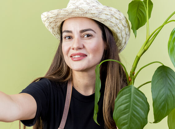 举行微笑美丽的园丁女孩穿着制服戴着园艺帽子拿着植物与相机隔离在橄榄绿园艺制服橄榄色