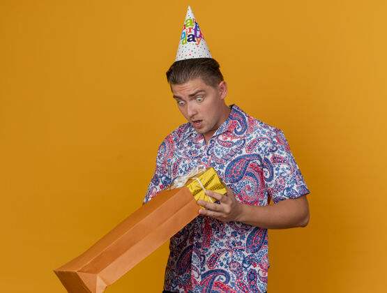 派对惊讶的年轻人戴着生日帽 拿着礼物袋看着孤立的橙色生日年轻人帽子