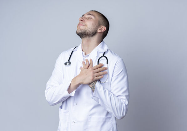 医学高兴的年轻男医生穿着医用长袍 脖子上戴着听诊器 双手放在心脏上 闭着眼睛隔离在白墙上周围姿势医生