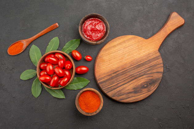 餐具俯视图一碗樱桃番茄 在黑桌子上放一个砧板 一个木勺 几碗番茄酱和辣椒粉黑食物碗