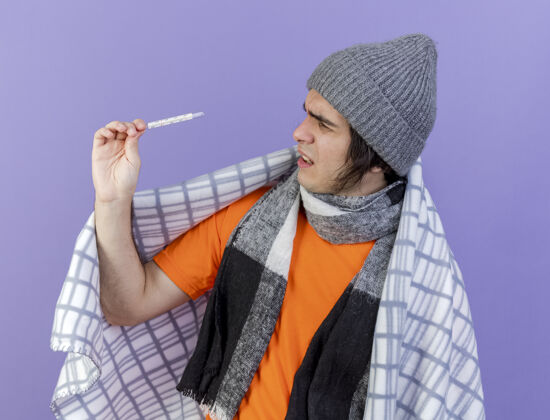 迷糊困惑的年轻病人戴着冬天的帽子 围巾裹着格子布 拿着温度计看着隔离在紫色上戴着抱着男人