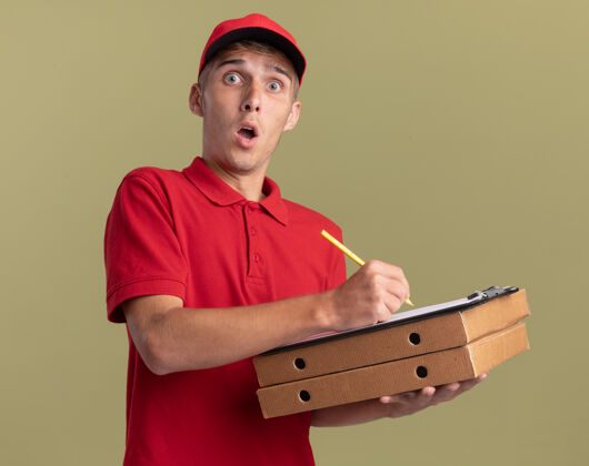 盒子惊讶的年轻金发送货员拿着铅笔和剪贴板在橄榄绿的比萨饼盒上惊喜剪贴板男孩