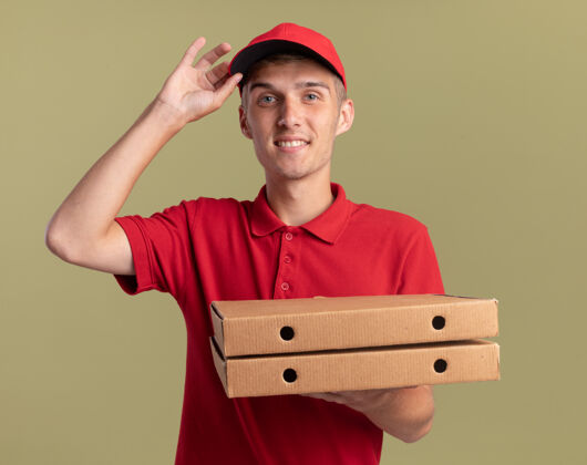 手微笑的年轻金发送货男孩把手放在帽子上 拿着橄榄绿的比萨饼盒金发年轻盒子