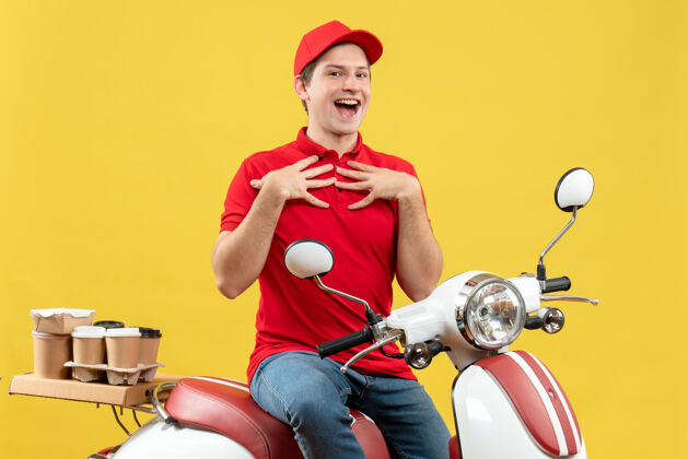 年轻人俯视图：身穿红色上衣 头戴帽子的年轻人坐在滑板车上 指着黄色的墙上 微笑着传递着命令衬衫命令传送带