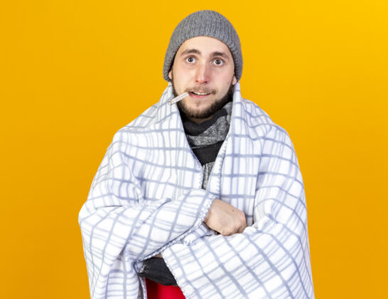 帽子兴奋的年轻白种人 戴着冬天的帽子 围着方格布围巾 把嘴里的温度计隔离在橙色的墙上 留着复印空间包裹男人穿