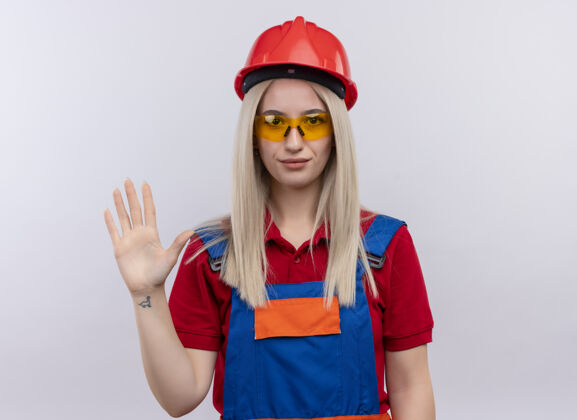 建筑工人一个穿着制服 戴着安全眼镜的年轻金发建筑工人女孩在与世隔绝的空白处打招呼年轻眼镜手势