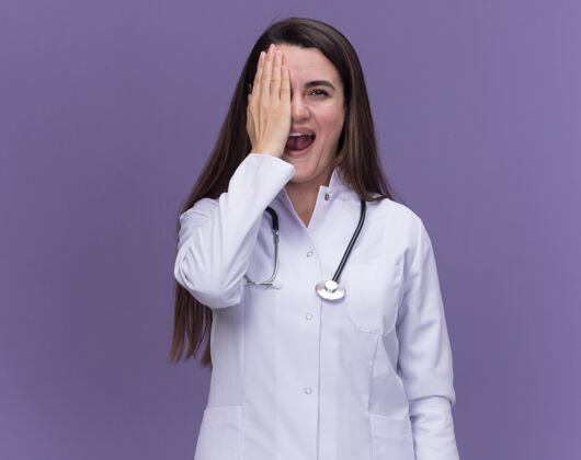 年轻快乐的年轻女医生穿着带听诊器的医用长袍 手捂着紫色的眼睛手听诊器医生