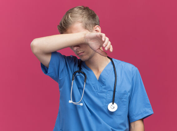 擦疲惫的年轻男医生穿着医生制服 用听诊器擦拭额头 手被隔离在粉红色的墙上人年轻人脸