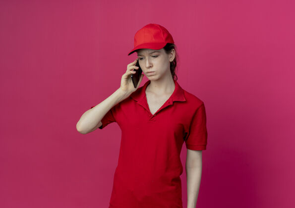 红色穿着红色制服 戴着帽子 正在打电话的年轻漂亮的送货女孩 在深红色背景下 带着复印空间 孤零零地往下看帽子羽绒服漂亮