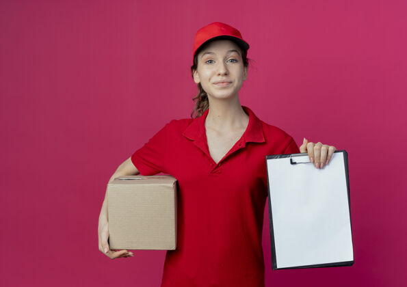 红色高兴的年轻漂亮的送货女孩穿着红色制服和帽子举行纸箱和剪贴板上的深红色背景与复制空间隔离背景年轻制服