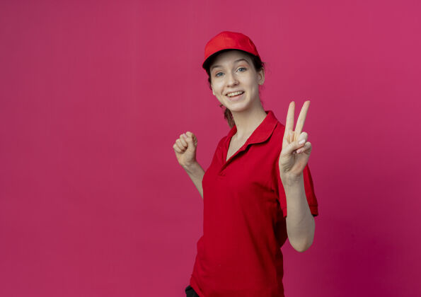 立场快乐的年轻漂亮的送货女孩穿着红色制服 戴着帽子站在侧视图中握紧拳头 做着和平的标志 在深红色的背景上与复制空间隔离制服女孩握紧