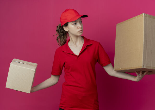 背景令人印象深刻的年轻漂亮的送货女孩在红色制服和帽子举行纸箱看着一侧孤立的深红色背景红色漂亮女孩