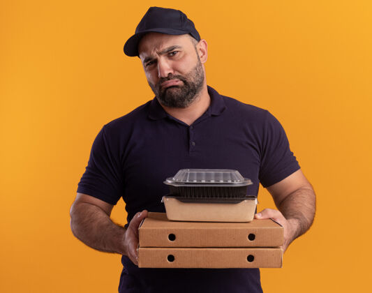 容器悲伤的中年送货员穿着制服 戴着帽子 拿着食物容器放在隔离在黄色墙上的比萨饼盒上制服衣服披萨