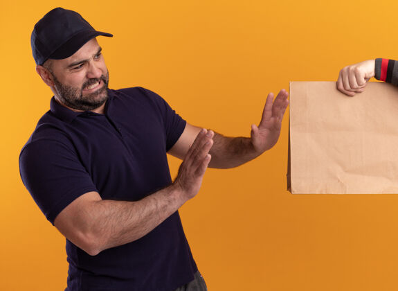 立场一个穿着制服 戴着帽子的中年送货员把纸食品包装交给隔离在黄色墙上的客户客户包装食物