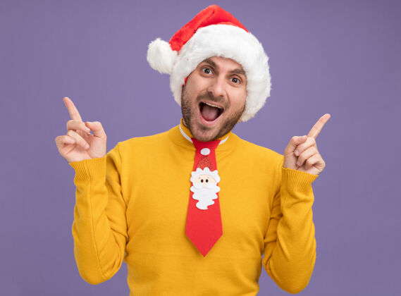 印象令人印象深刻的年轻白人男子戴着圣诞帽和领带看着摄像机指着孤立的紫色背景男人指向圣诞