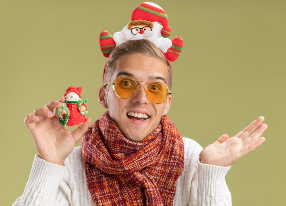 绿色印象深刻的年轻帅哥戴着圣诞老人的头带和围巾看着相机拿着雪人圣诞饰品显示空手孤立在橄榄绿的背景举行圣诞老人显示
