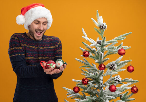 帽子令人印象深刻的年轻白种人戴着圣诞帽站在装饰圣诞树附近拿着圣诞饰品看橙色背景孤立男人橘子圣诞饰品