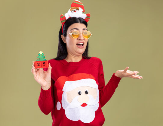 显示恼怒的年轻白人女孩戴着圣诞老人的头带和毛衣 戴着眼镜 手里拿着圣诞树玩具和日期 一边看一边显示空手孤立在橄榄绿的背景上眼镜头带圣诞树