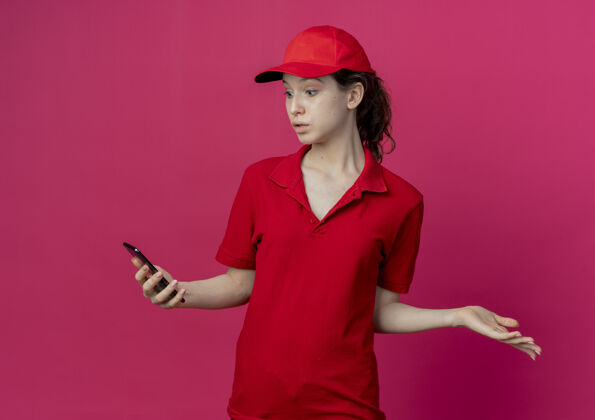 制服令人印象深刻的年轻漂亮的送货女孩在红色制服和帽子举行 看着手机和显示空的手孤立在深红色的背景手机红色送货