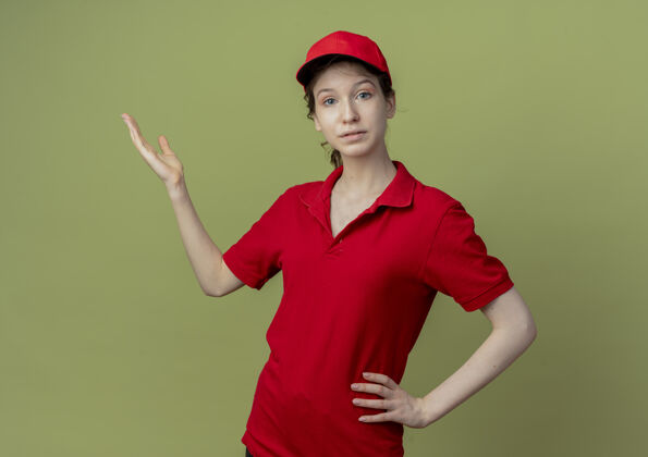 腰年轻漂亮的送货女孩 穿着红色制服 戴着帽子 看着相机 把手放在腰上 在橄榄绿的背景上 显示出一只空的手 还有复印空间帽子空橄榄