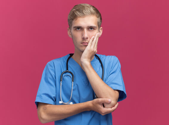 制服严格的年轻男性医生穿着医生制服 用听诊器将手放在脸颊上 隔离在粉红色的墙上 留有复印空间感情脸听诊器