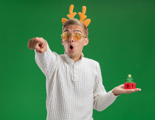 鹿角令人印象深刻的年轻帅哥戴着驯鹿鹿角头带戴着眼镜拿着圣诞树玩具和日期看 并指着绿色背景上孤立的一面圣诞快乐举行年轻人