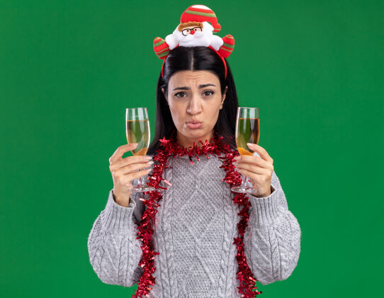 香槟困惑的年轻白人女孩戴着圣诞老人的头带 脖子上戴着金箔花环 手里拿着两杯香槟 看着隔离在绿色背景上的相机圣诞老人头带新年