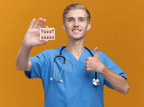 制服微笑着的年轻男医生穿着医生制服手持听诊器的药片 在橙色的墙上孤立地竖起大拇指站着抱着表演