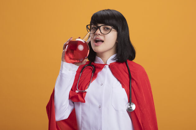 人穿着医用长袍听诊器和带眼镜斗篷的年轻超级英雄女孩高兴地喝着化学玻璃里的红色液体医学持有红色