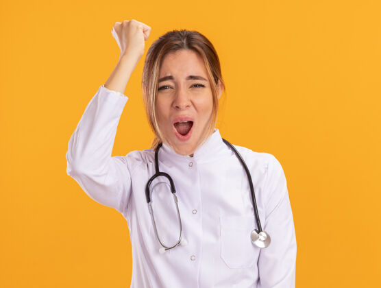人快乐的年轻女医生穿着医用长袍 手持听诊器举起拳头隔离在黄色的墙上女性听诊器医生