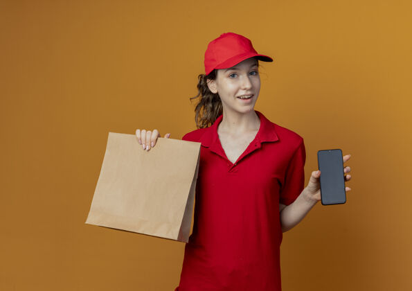 帽子令人印象深刻的年轻漂亮的送货女孩在红色制服和帽子显示纸包装和手机在橙色的背景与复制空间隔离送货手机年轻