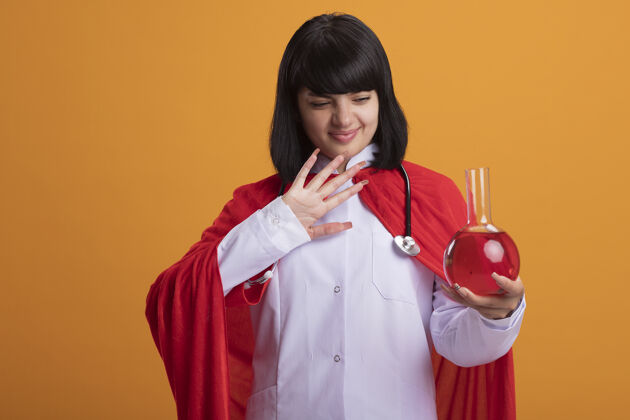 穿未被释放的年轻超级英雄女孩戴着听诊器 穿着医用长袍和斗篷 拿着化学玻璃瓶 里面装满了隔离在橙色上的红色液体橙色年轻瓶子
