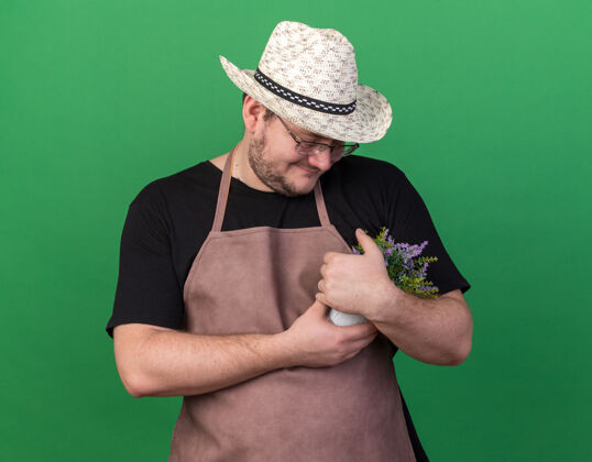 帽子高兴的年轻男园丁戴着园艺帽抓起花盆 看着隔离在绿色墙上的花朵请花花盆