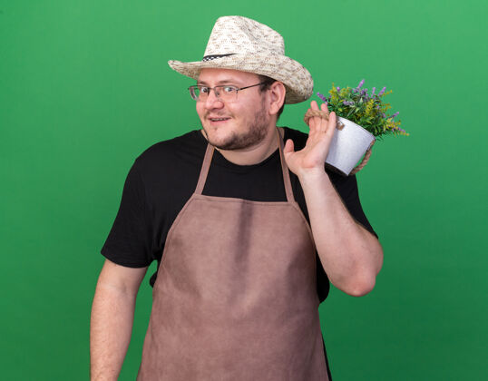 男性微笑着的年轻男园丁戴着园艺帽 肩上扛着花盆里的花 隔离在绿色的墙上花盆花年轻