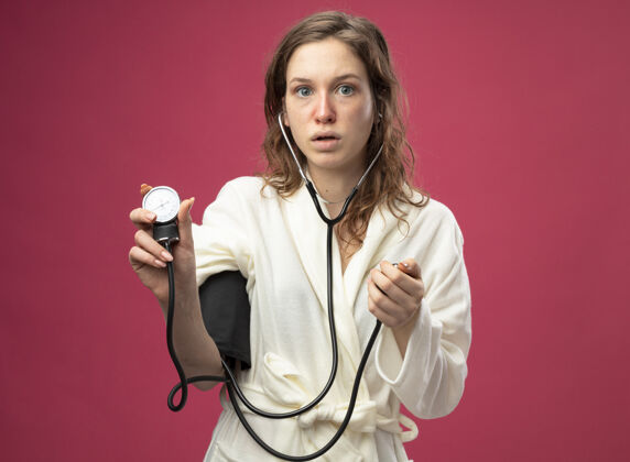 惊讶惊讶的年轻病女孩穿着白色长袍测量自己的压力血压计隔离在粉红色疾病测量血压计