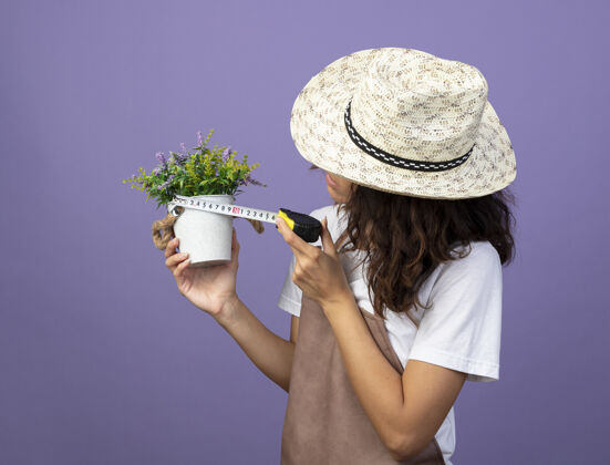 女人穿着制服的沉思的年轻女园丁戴着园艺帽 用卷尺在紫色的花盆里量着花年轻
