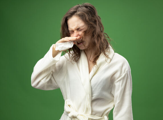 长袍未出狱的年轻病女孩闭着眼睛穿着白色长袍擦鼻子用餐巾隔离在绿色餐巾年轻绿色