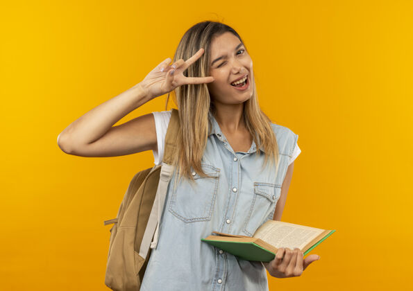 和平快乐的年轻漂亮的学生女孩戴着背袋拿着打开的书眨眼做和平标志孤立在橙色高兴年轻抱