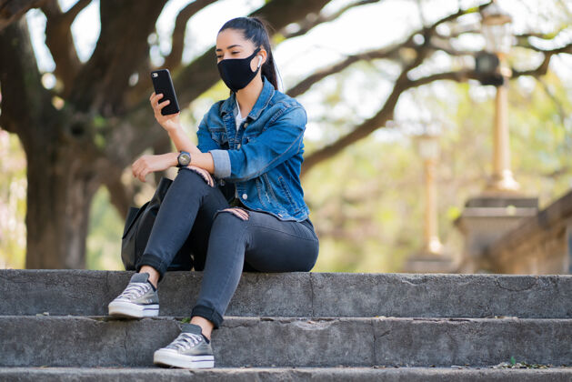 移动年轻女子戴着口罩 坐在室外楼梯上使用手机的画像城市放松年轻